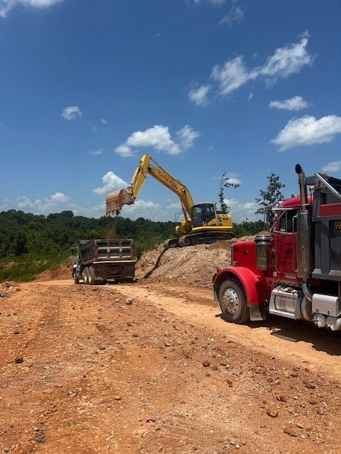 Yellow excavator loading dirt into two dump trucks on a dirt road. Bright sky, trees in background.