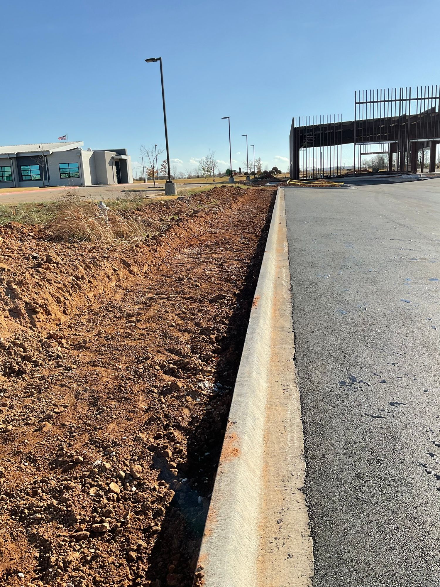 Dirt border alongside asphalt road and curb, with buildings in background under clear sky.
