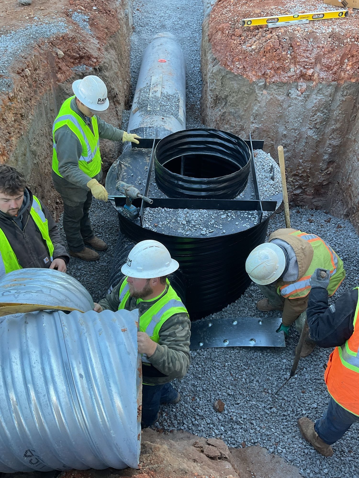 Construction workers installing a corrugated metal culvert and manhole in a trench, gravel base.