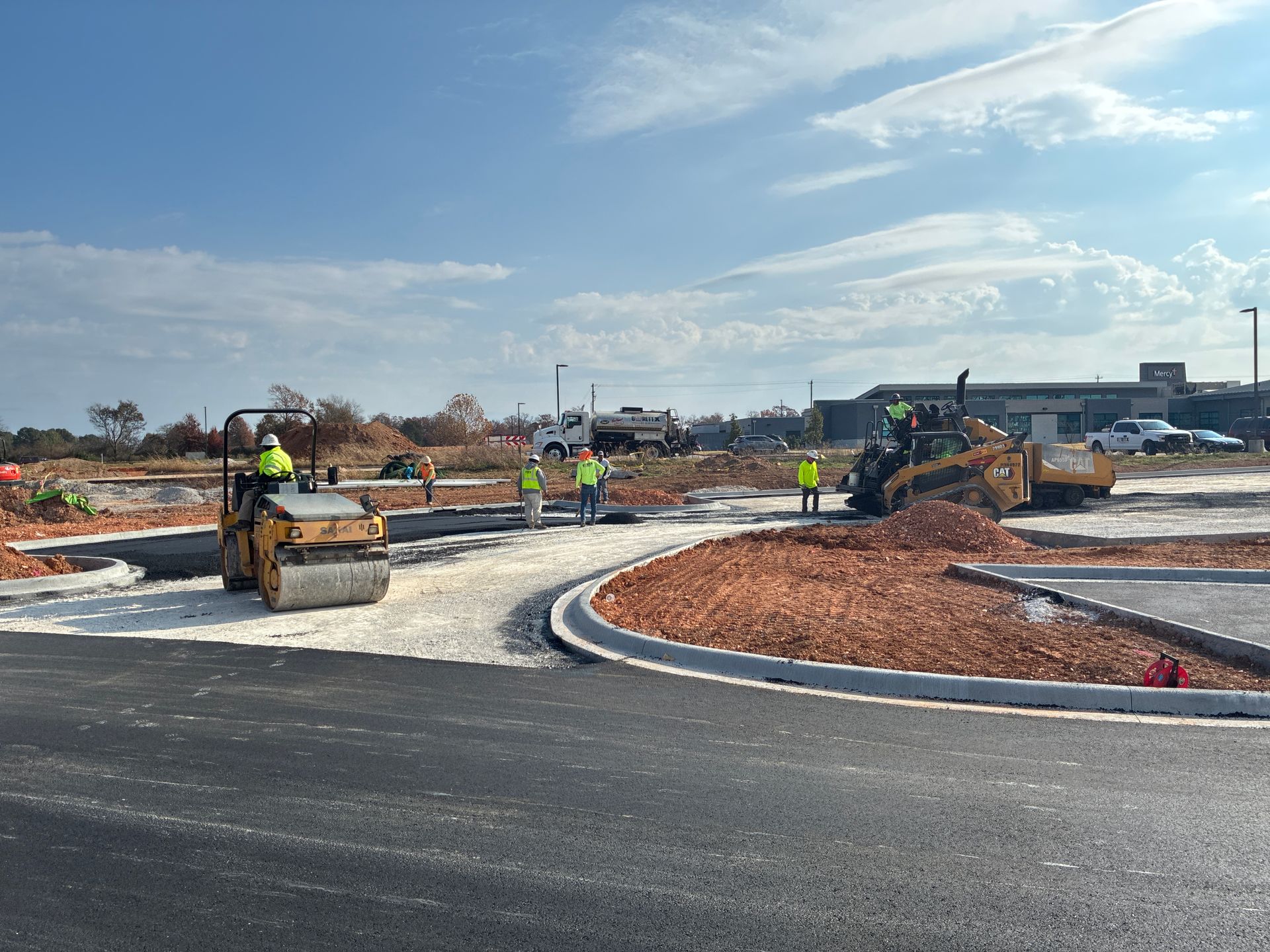 Construction site with asphalt and gravel paving, workers, and heavy machinery under a cloudy sky.