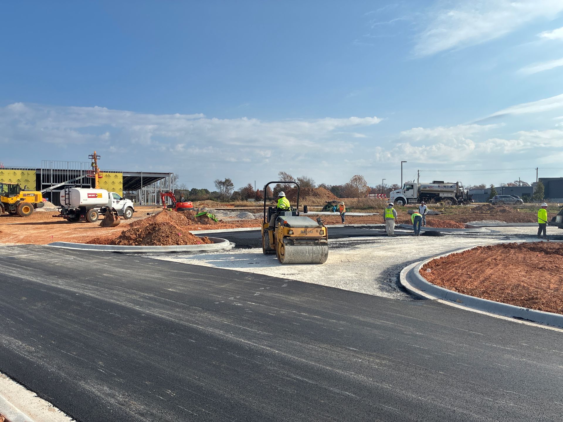Construction site with workers, a road roller compacting gravel, and a partially paved road.