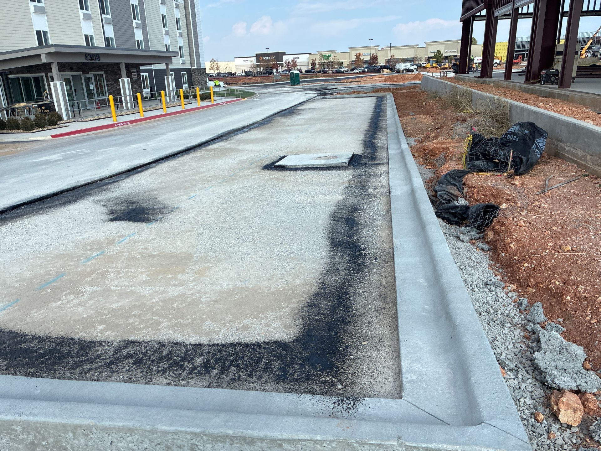 Construction site with gravel and asphalt surfaces next to a concrete curb. A hotel is visible in the background.