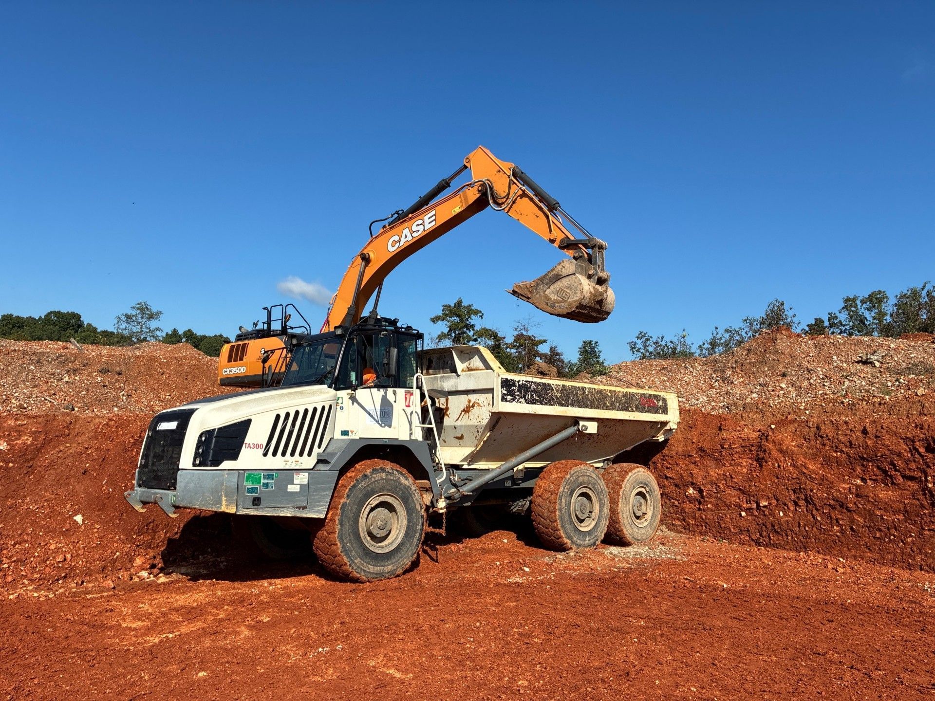 Excavator loading dirt into a large, white dump truck in a red dirt quarry under a blue sky.