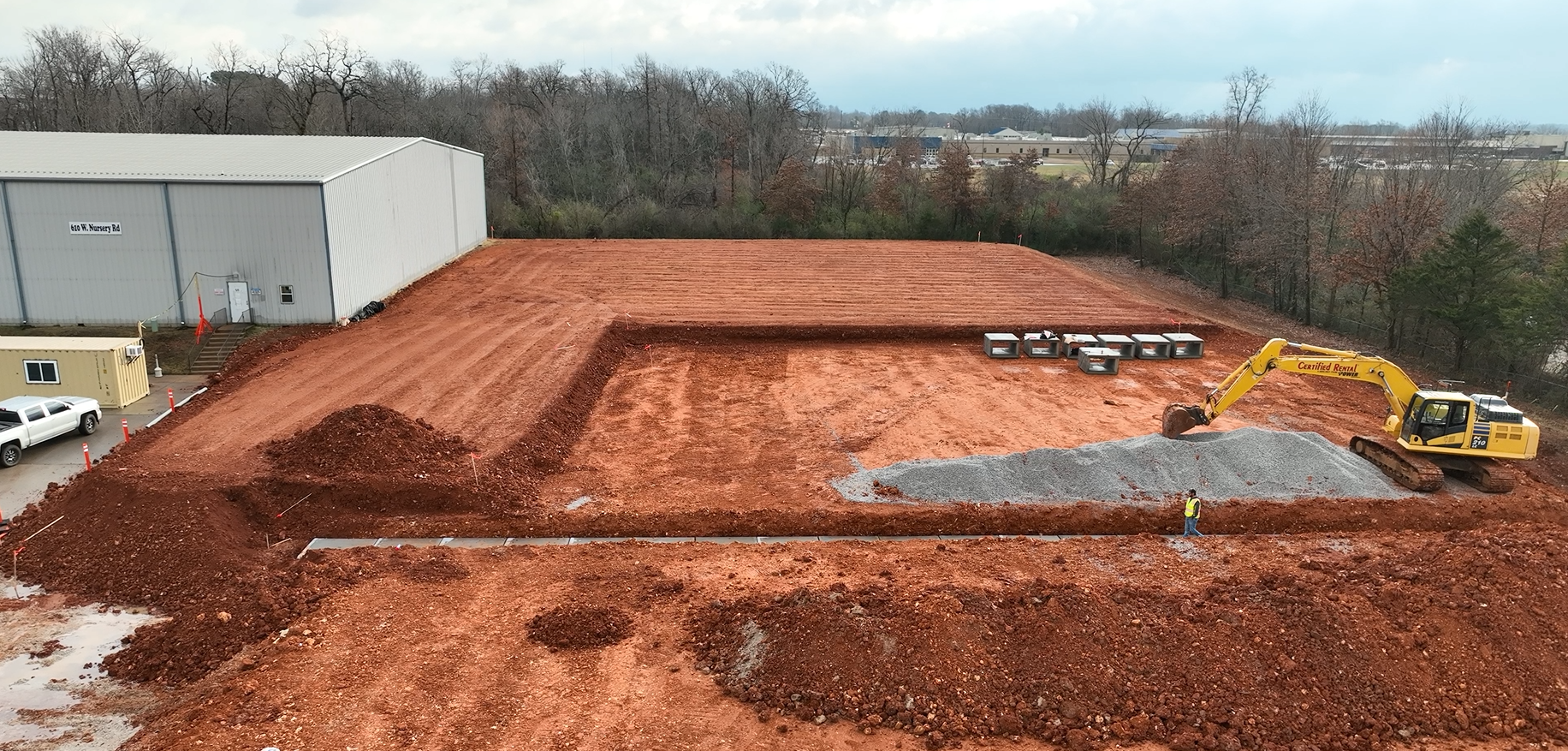 Construction site with an excavator moving gravel. Red dirt surrounds a warehouse.