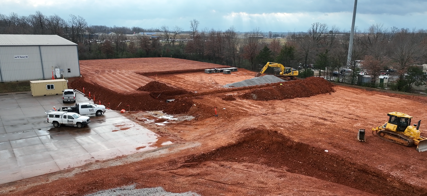 Construction site with dirt piles, heavy machinery, and work trucks. Gray warehouse and cloudy sky in background.