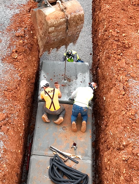 Construction workers installing concrete block in a trench. An excavator bucket hangs above them.