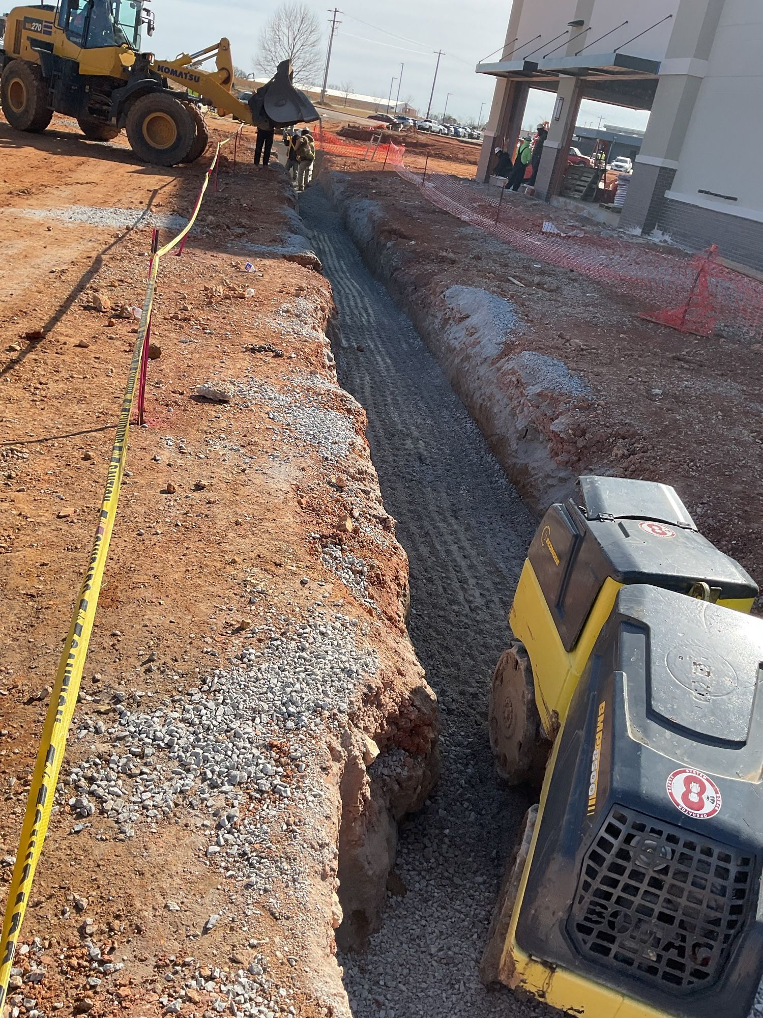 Construction site: Excavated trench with gravel, a compacting machine, and a front-end loader.