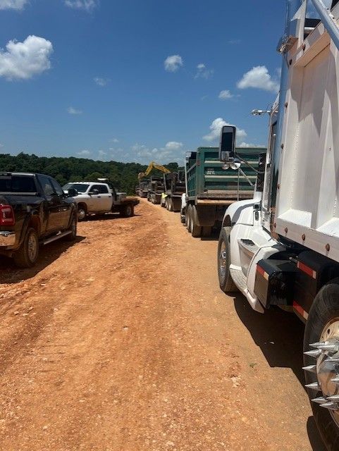 Line of construction vehicles on a red dirt road under a blue sky.
