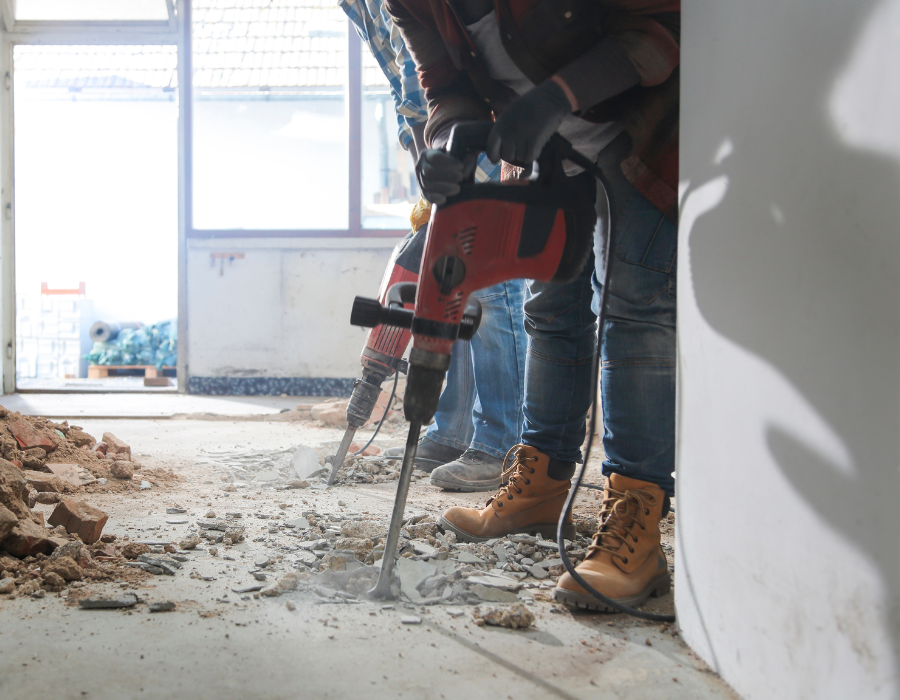 Gli operai utilizzano martelli pneumatici per rompere il pavimento di cemento in una stanza in fase di ristrutturazione, indossando stivali da lavoro e guanti.