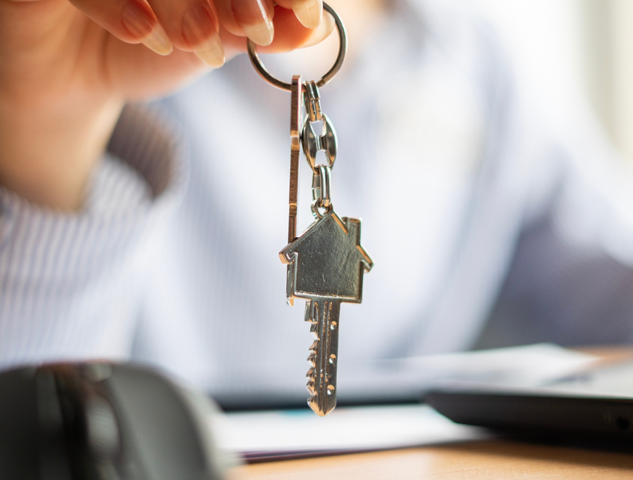 Mano di una persona che tiene le chiavi con un portachiavi a forma di casa, di fronte a una scrivania sfocata.