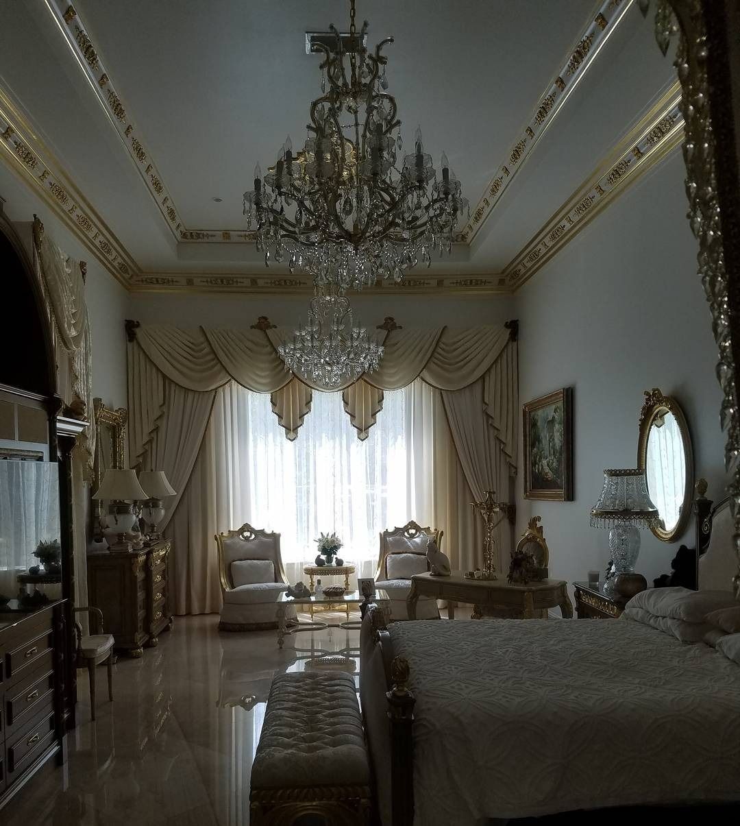 Opulent bedroom with chandelier, ornate gold accents, and large window with draped curtains.