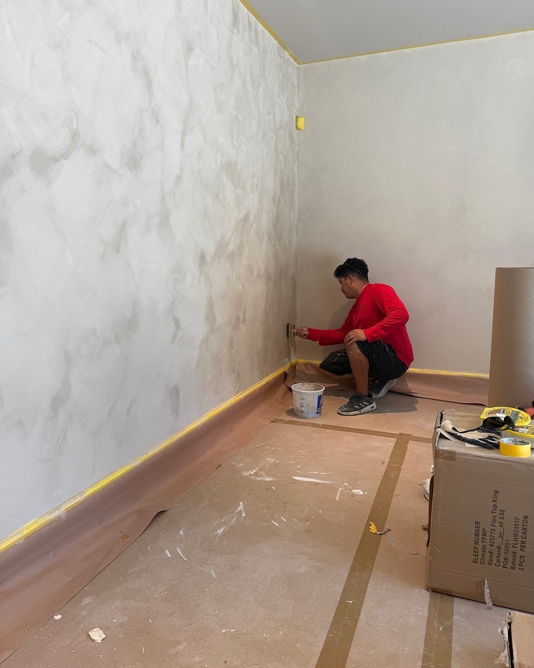 Man applying decorative paint on a wall, kneeling in a room with protective tape and cardboard.