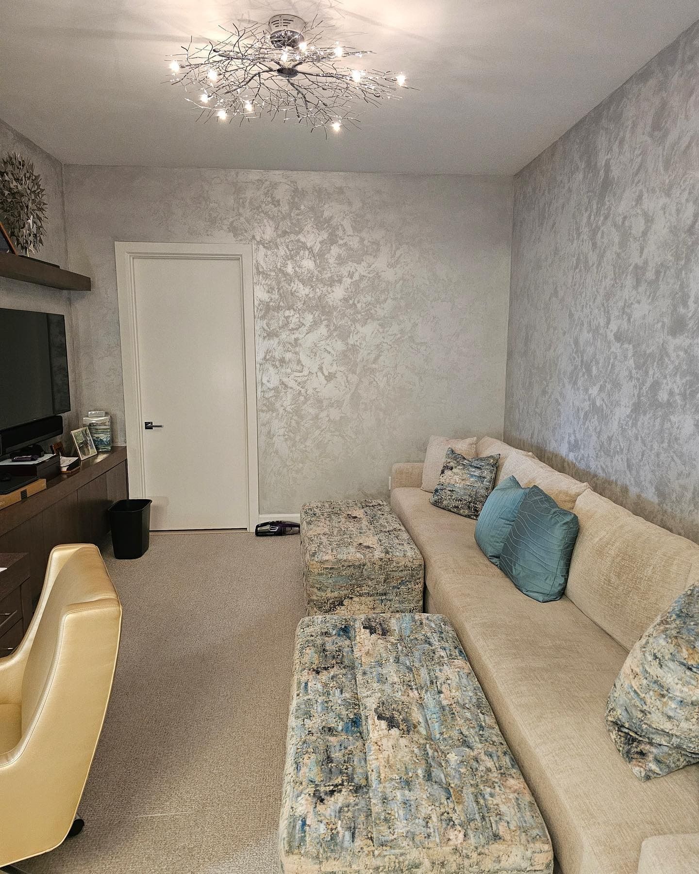 A living room with a beige couch, patterned ottomans, textured silver walls, and a decorative ceiling light.
