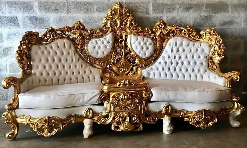 Gold-framed, ornate white loveseat with tufted back against a brick wall.