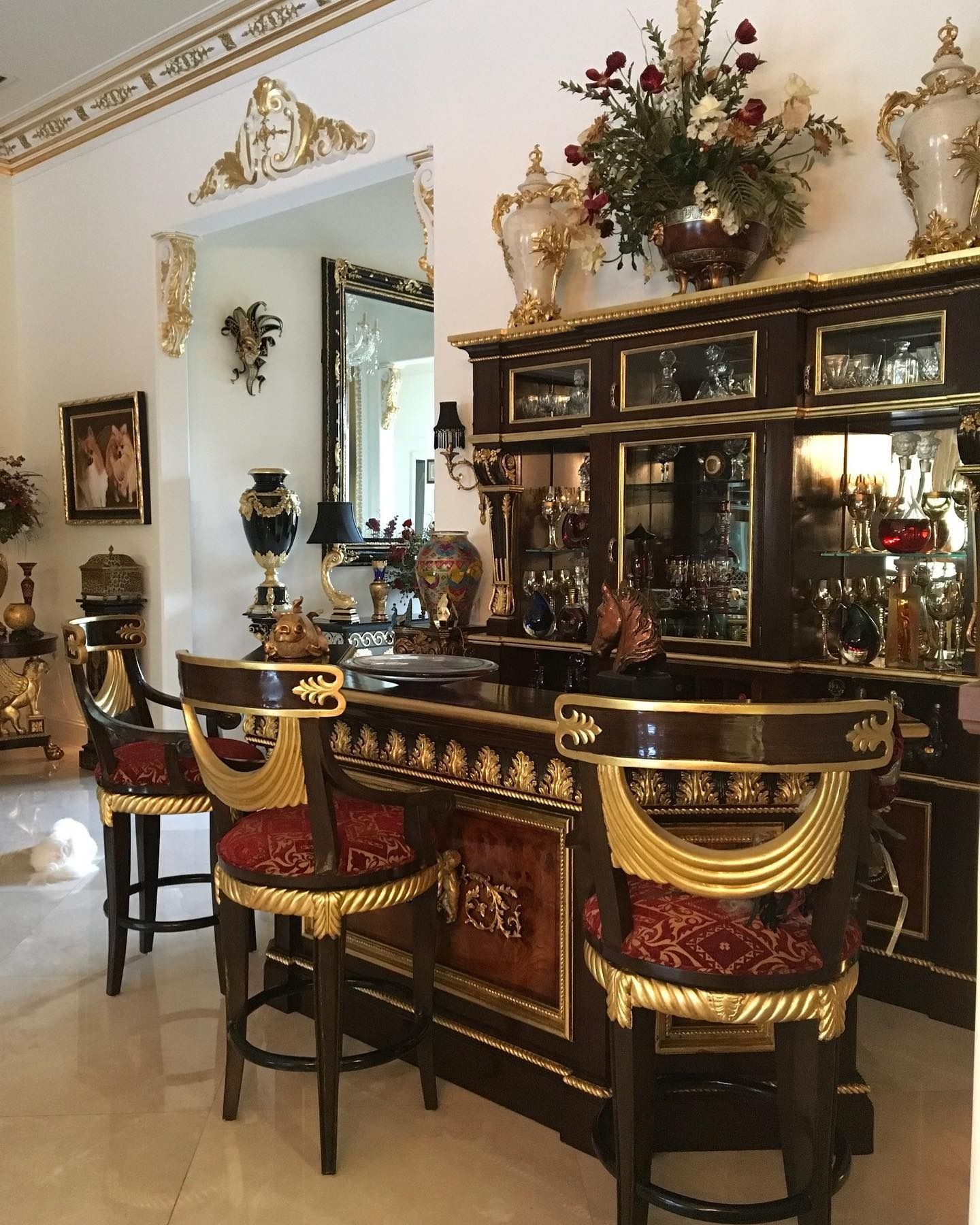 Ornate bar with gold accents, dark wood, and red upholstered stools in a bright, decorated interior.