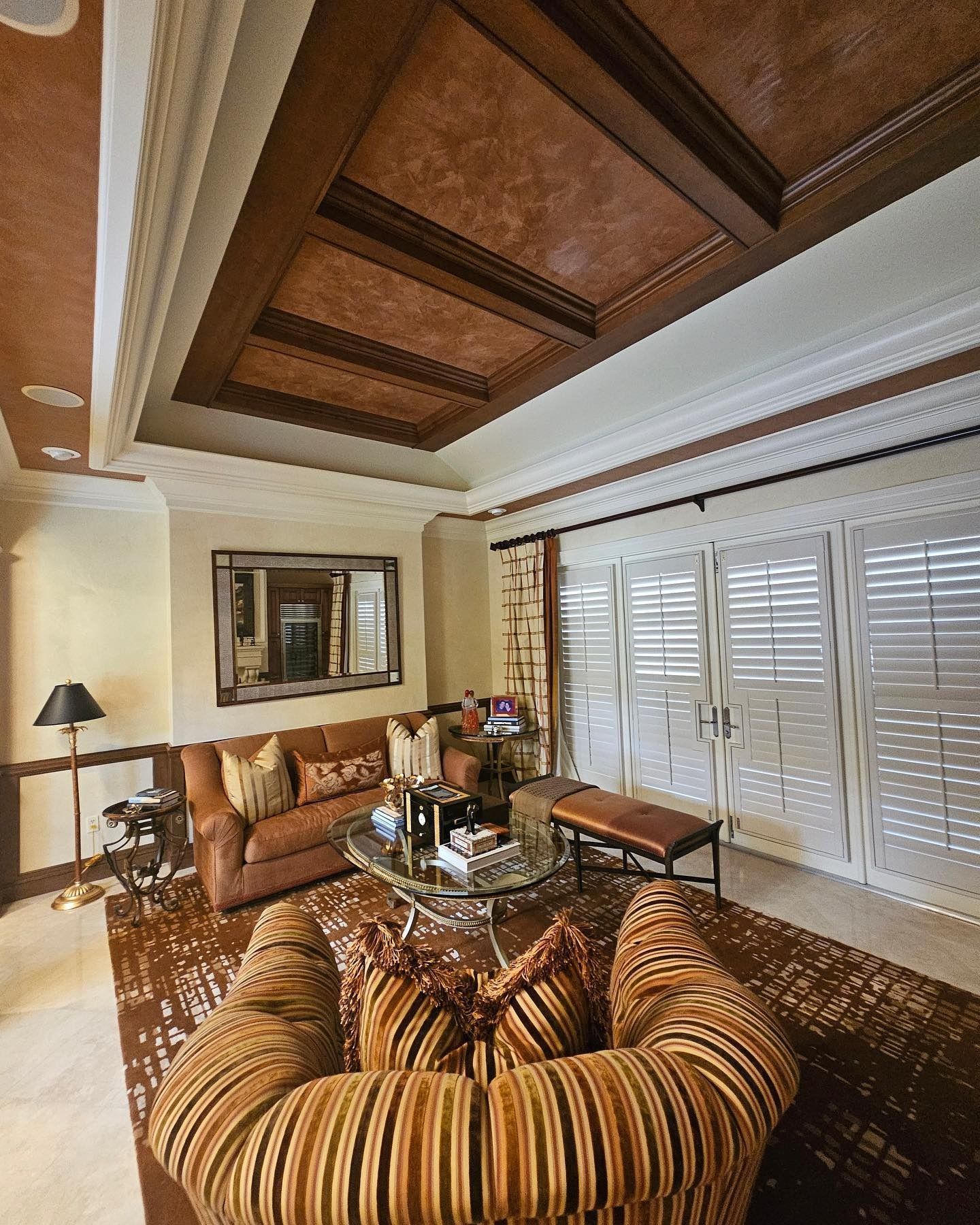 Living room with brown and gold decor: sofa, patterned chair, shutters, and coffered ceiling.