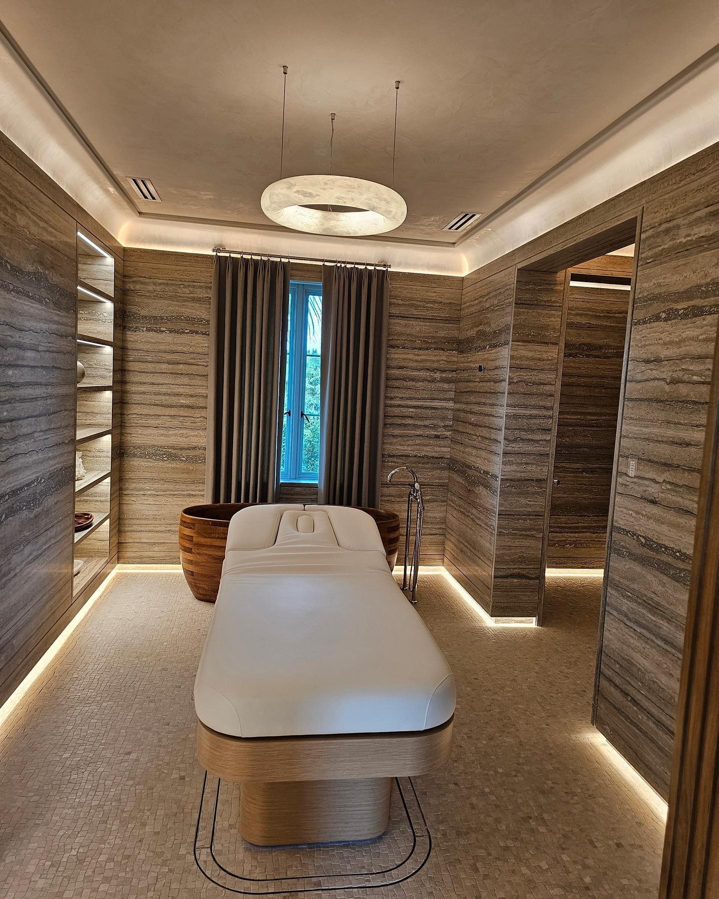Spa treatment room: massage table, stone walls, neutral tones, overhead lighting, window with curtains.