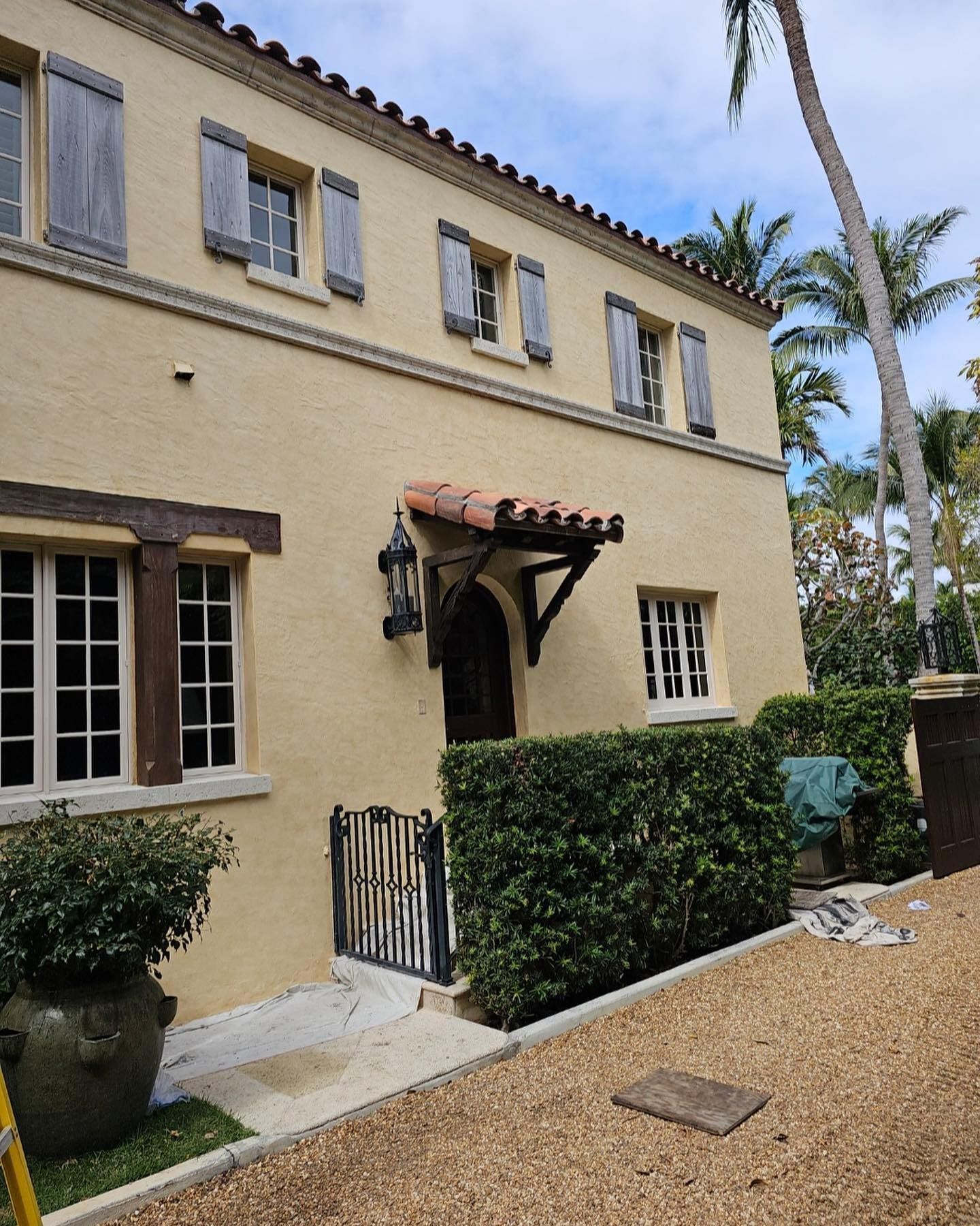 Beige stucco home with brown trim, shutters, and a tiled awning; shrubbery and a gravel path.