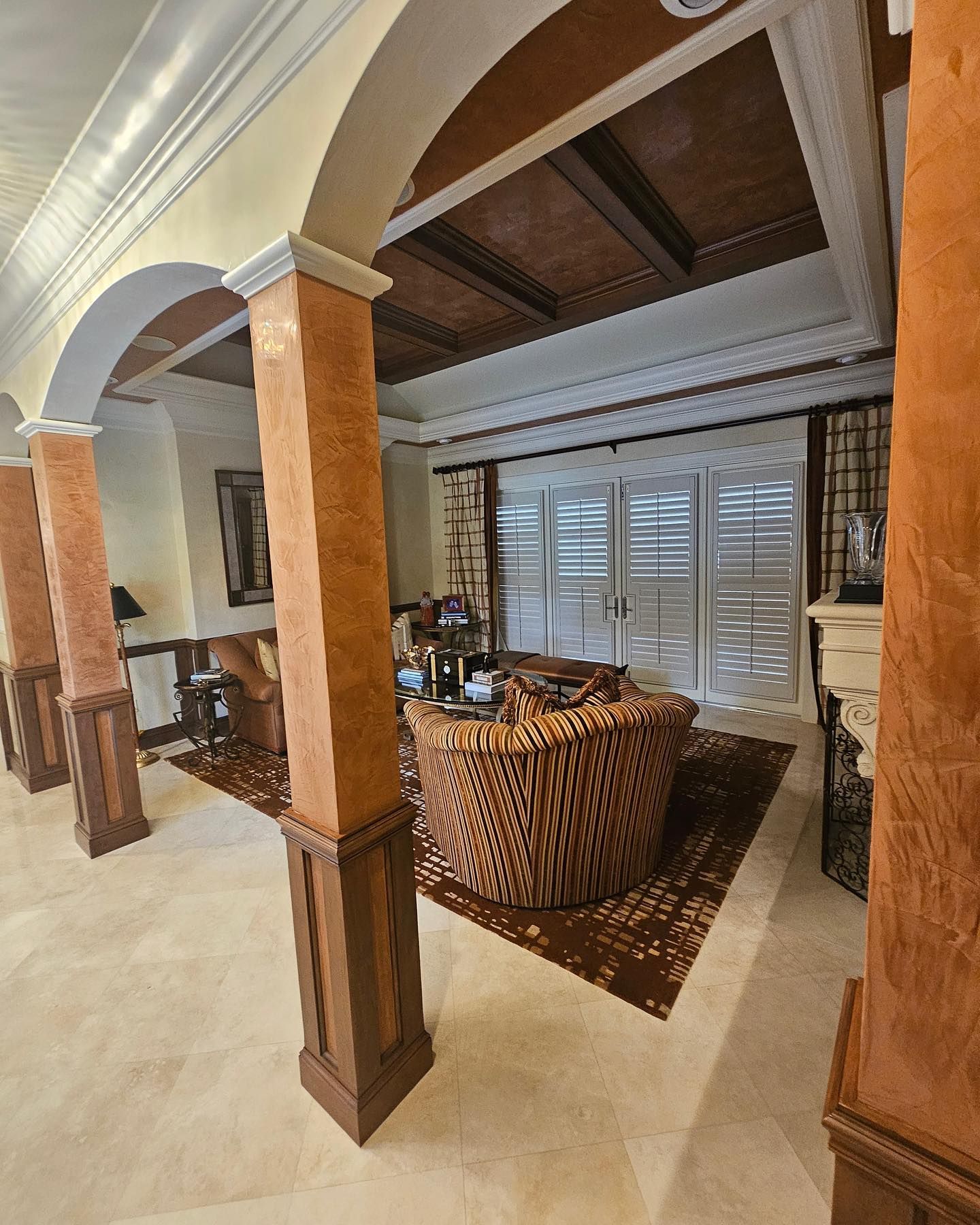 Living room with columns, brown seating, rug, and shuttered windows. Earth tones and classic architecture.