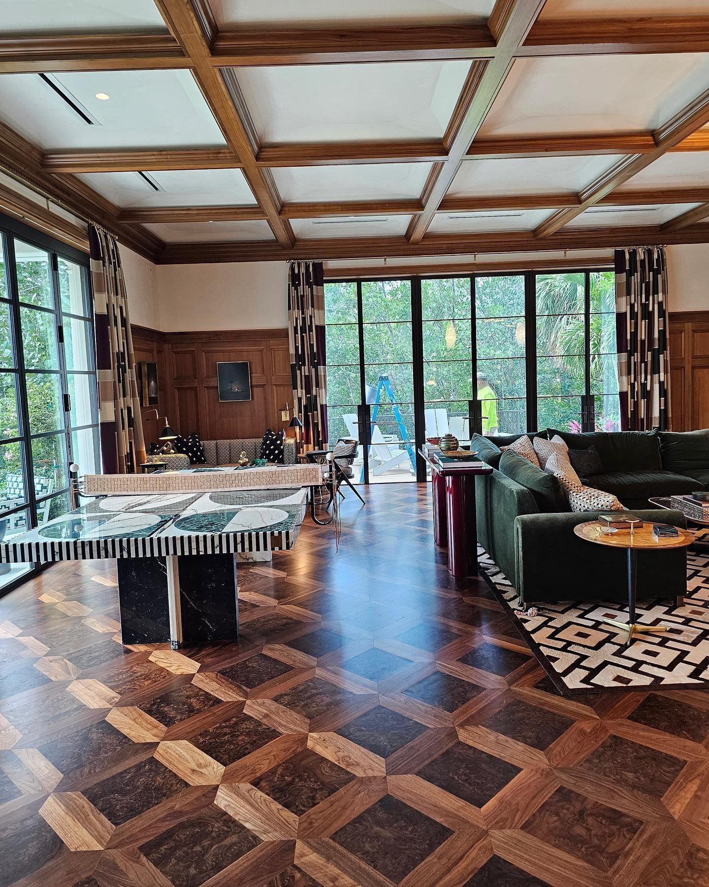 Luxurious living room: dark wood floors, coffered ceiling, large windows, green sofa, patterned rug, and a modern table.
