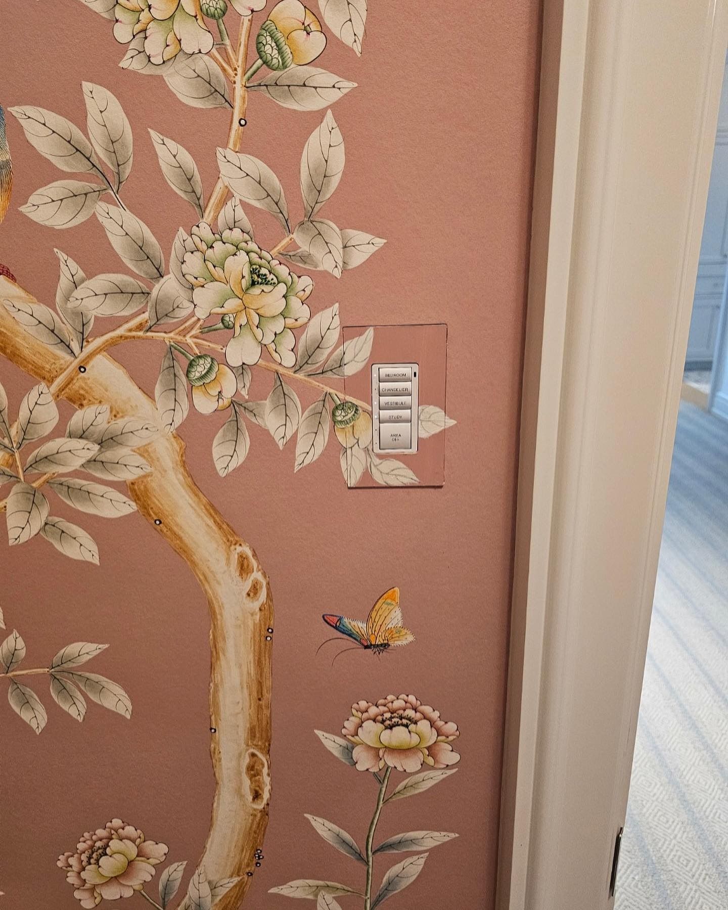 Wallpaper with a floral design, including a butterfly, surrounds a light switch on a pink wall.