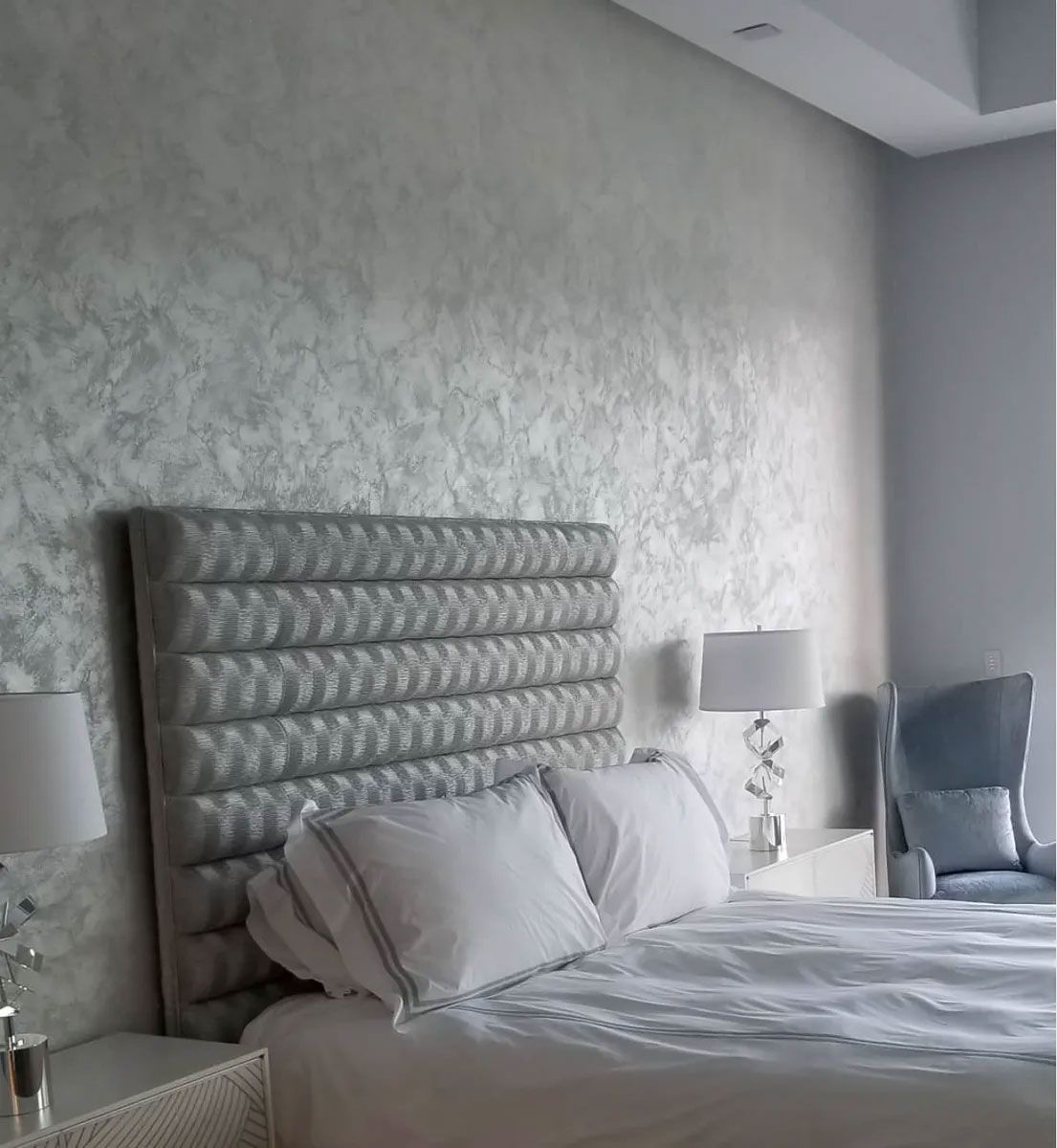 Bedroom with a bed, plush headboard, textured wall, bedside lamps, and a chair.