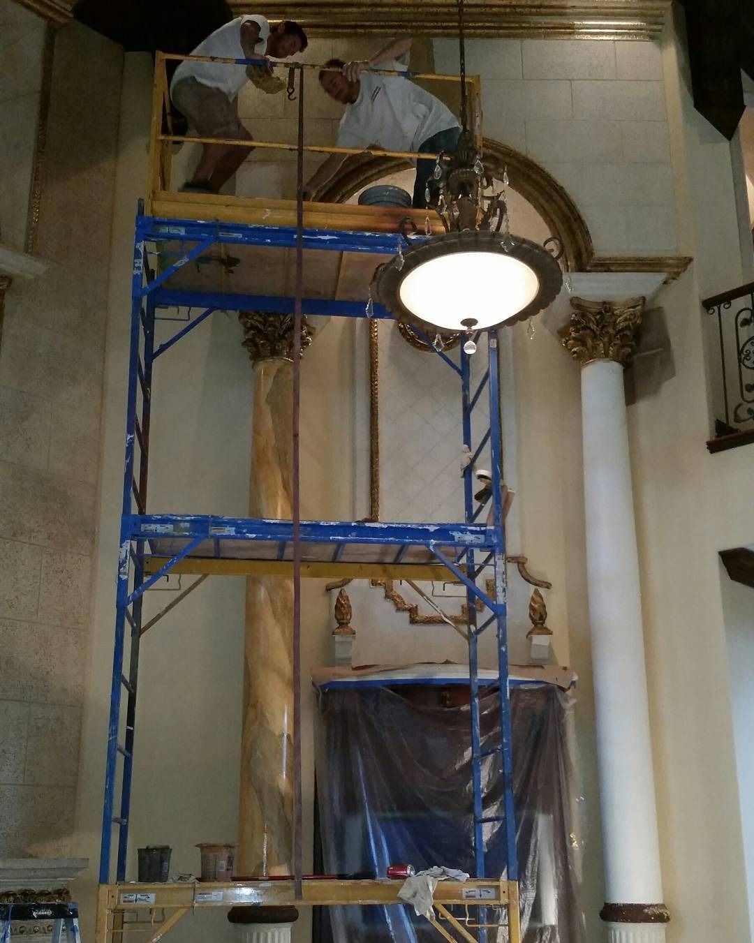 Two workers on a scaffold restoring ornate ceiling detail near a light fixture, likely inside a building.