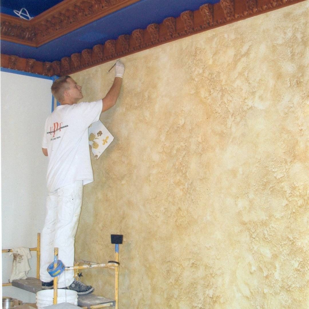A painter on a scaffold applies decorative finish to a wall with a brush; gold tones.