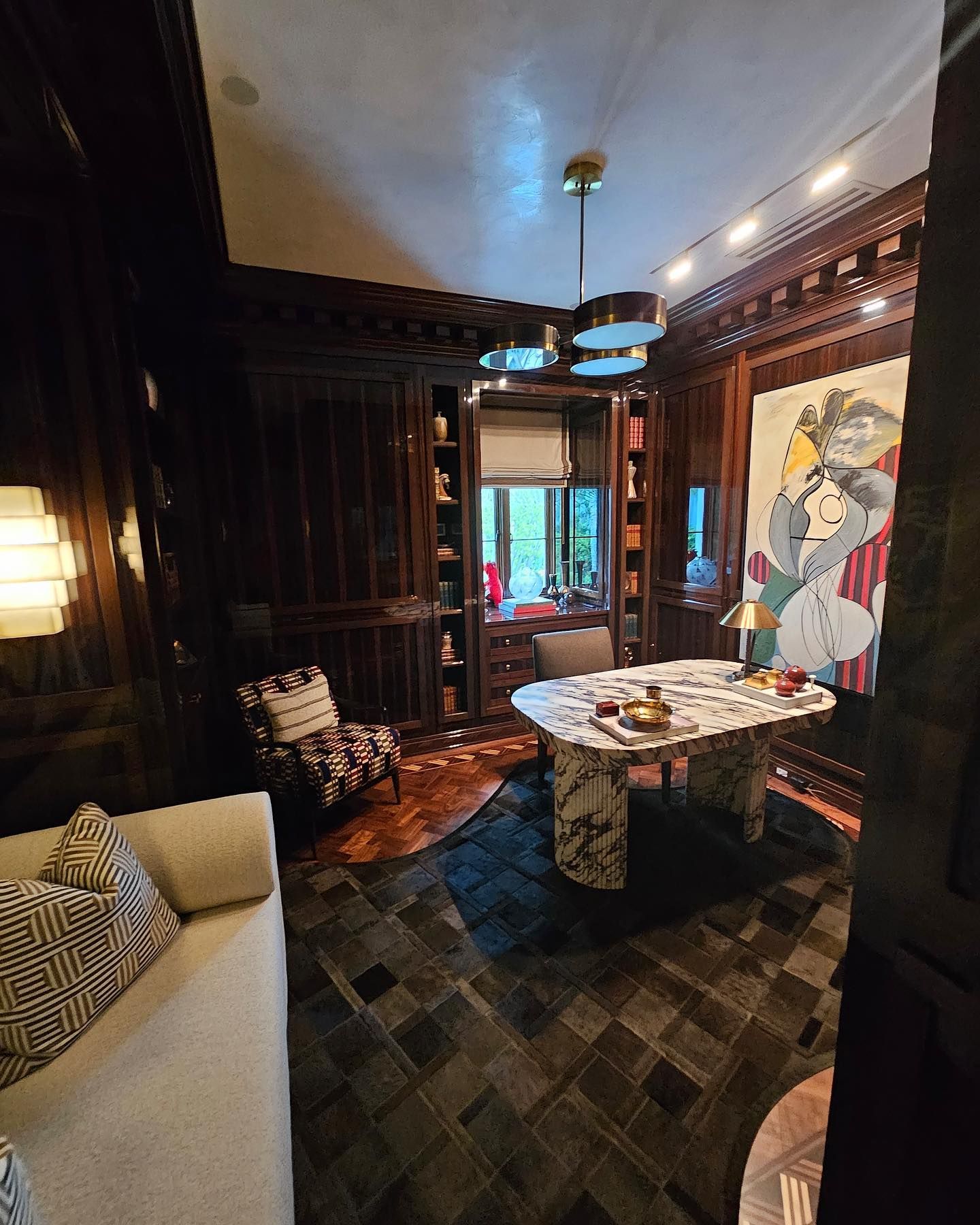 Luxurious, dark-paneled home office with art, marble desk, patterned rug, and accent chair.