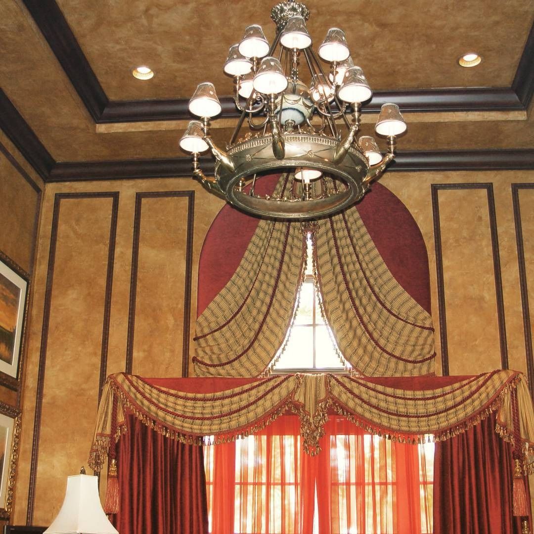 Ornate chandelier hangs above arched window draped with red and gold curtains in a luxurious room.