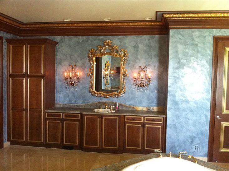 Bathroom with ornate gold-framed mirror, brown cabinets, blue marbled walls, and crystal sconces.