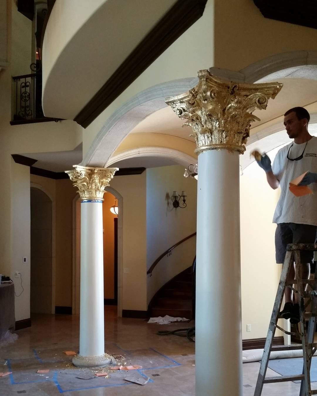 Man on a ladder gilding the gold capital of a pillar inside a building.