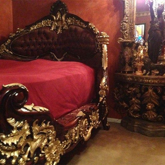 Ornate bed with gold accents and a red comforter in a room with a matching nightstand.