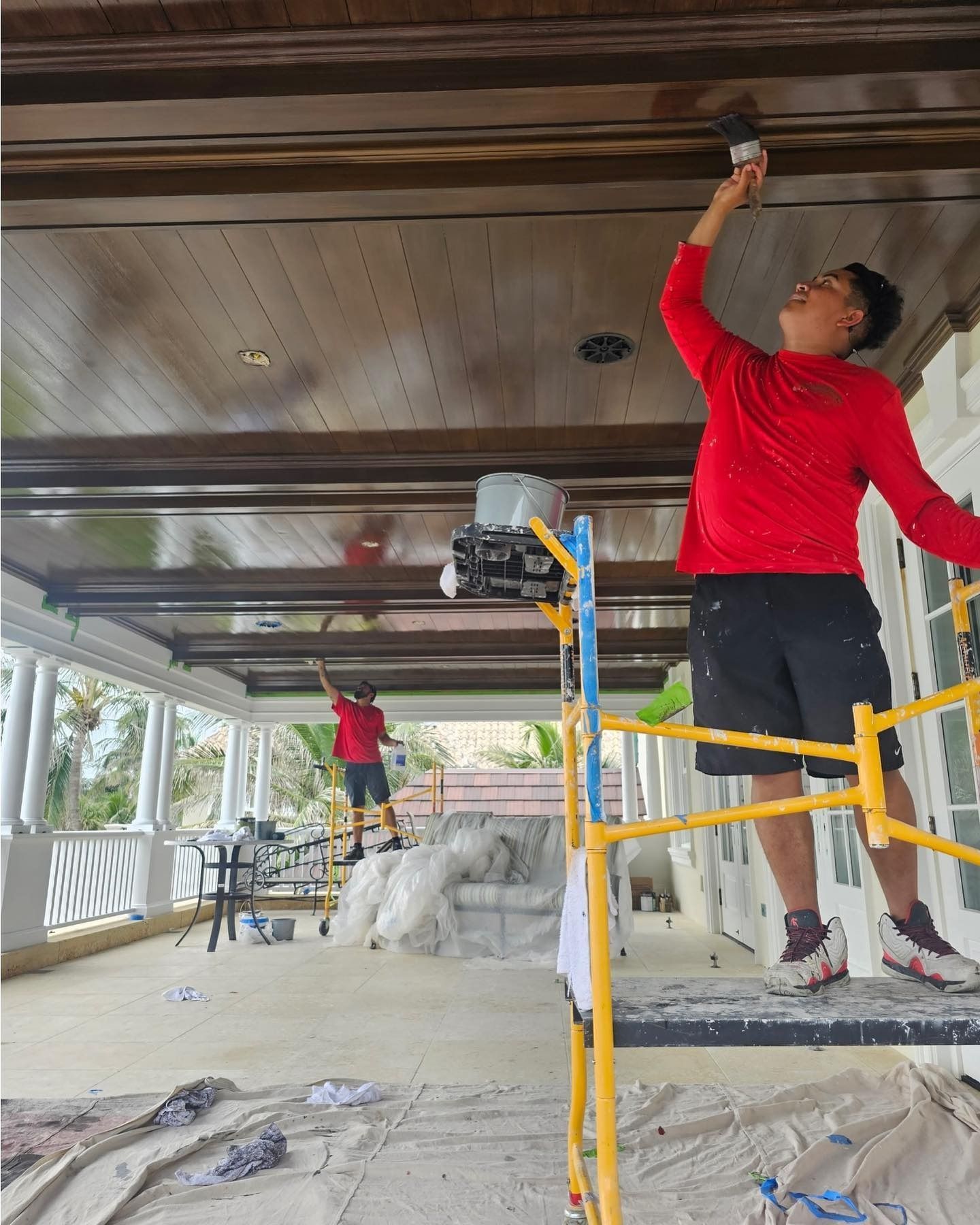 Two people painting a brown ceiling on a porch; one on a scaffolding, the other on a ladder.
