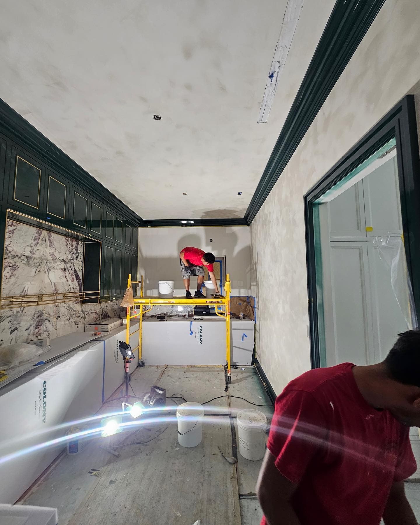 Two workers paint a room with green trim, one on scaffolding. Beige walls.