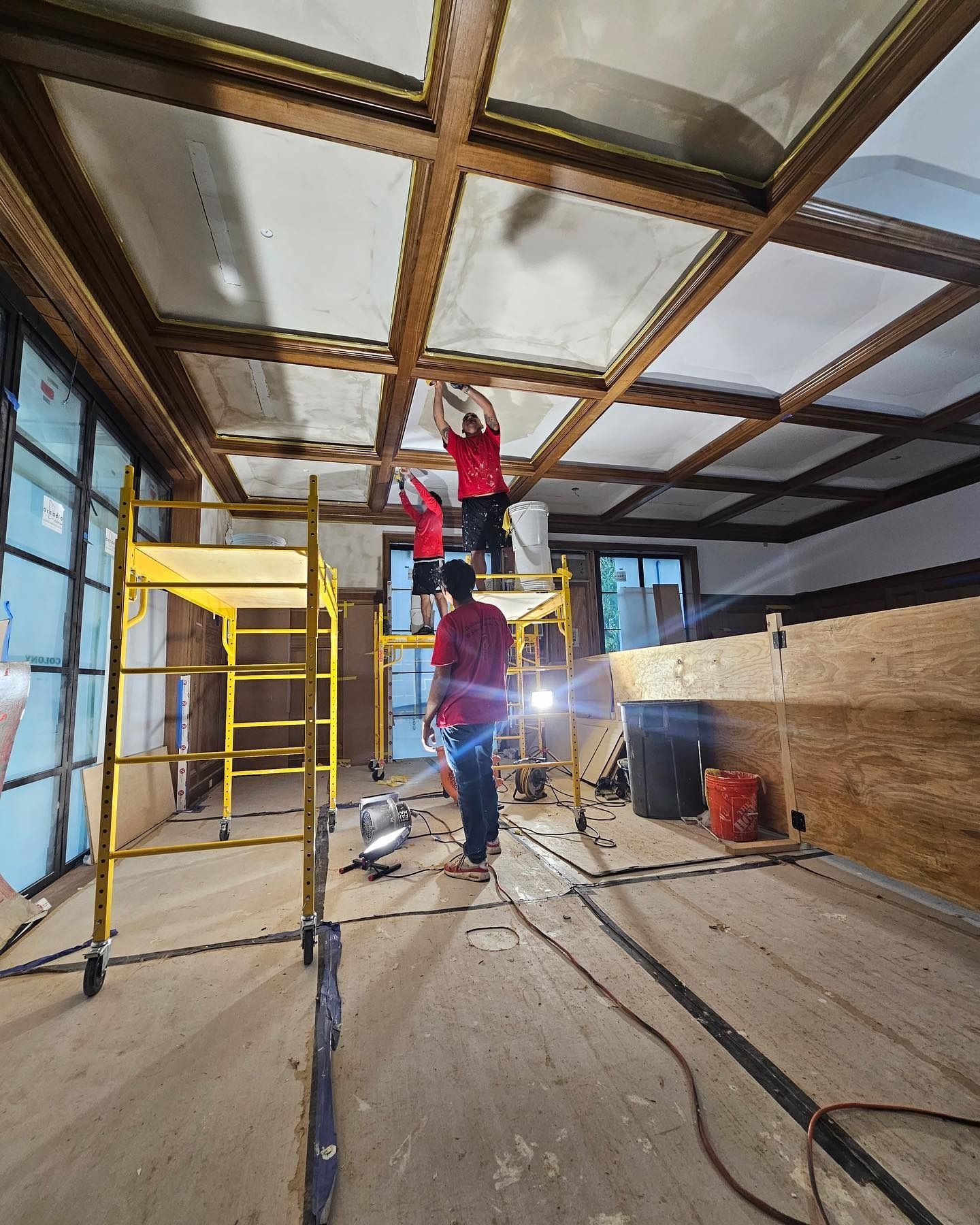 Construction workers installing ceiling panels in a room with scaffolding. Wooden beams and a light are visible.