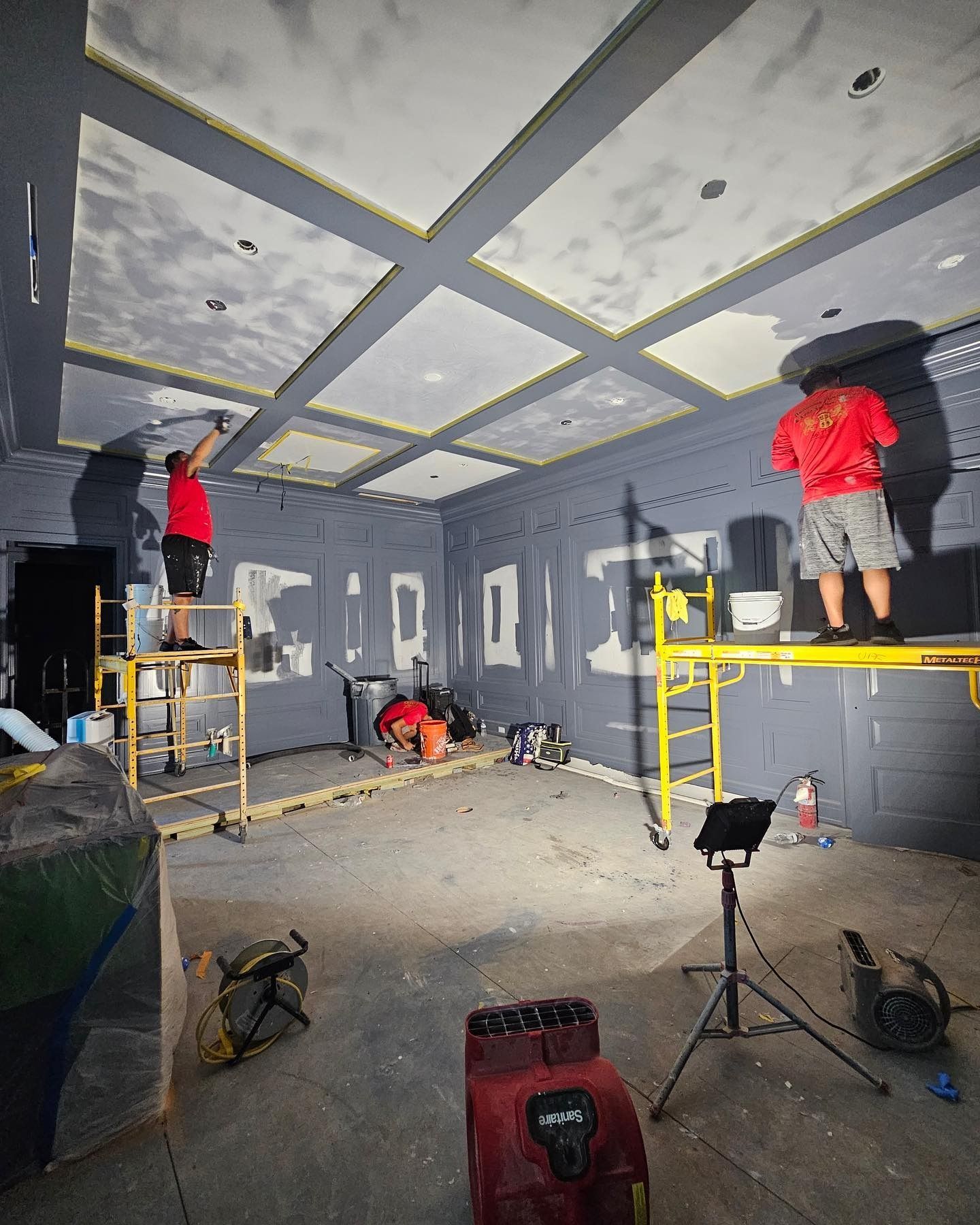 Two workers painting a room with a patterned ceiling and gray walls, standing on scaffolding.