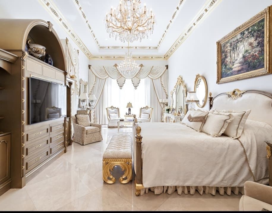 Luxurious bedroom with ornate gold and white furniture, chandelier, and artwork.
