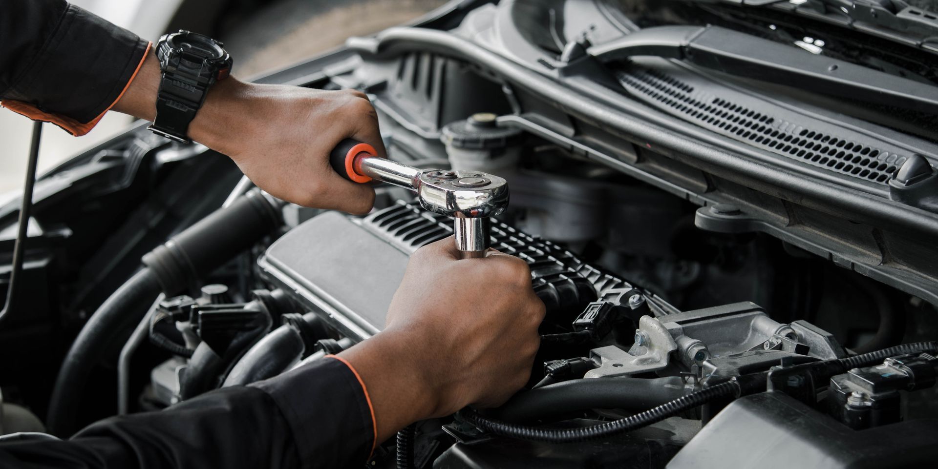 A mechanic working on a car engine with a wrench. The mechanic has dark skin and is wearing a black watch and shirt.