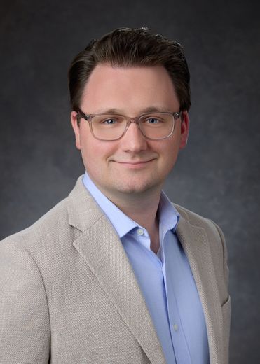 A man in a suit and a purple shirt is standing in front of a yellow background.