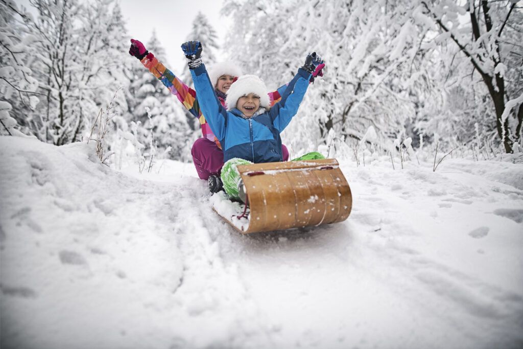 Two children are sledding down a snow covered hill.