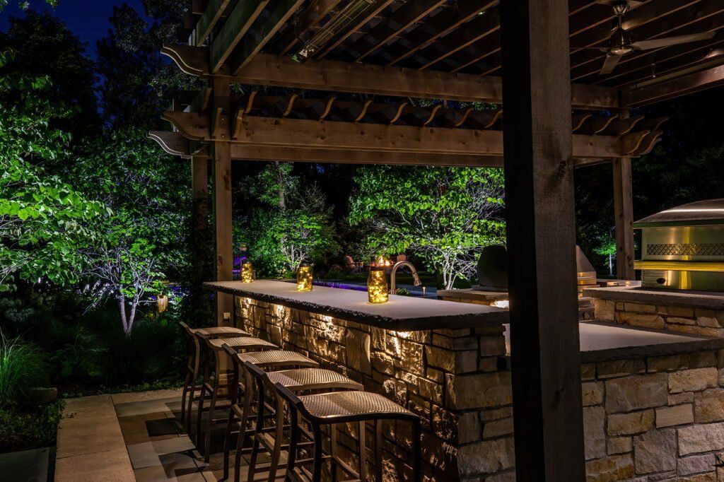 A patio with a bar and stools under a pergola at night.