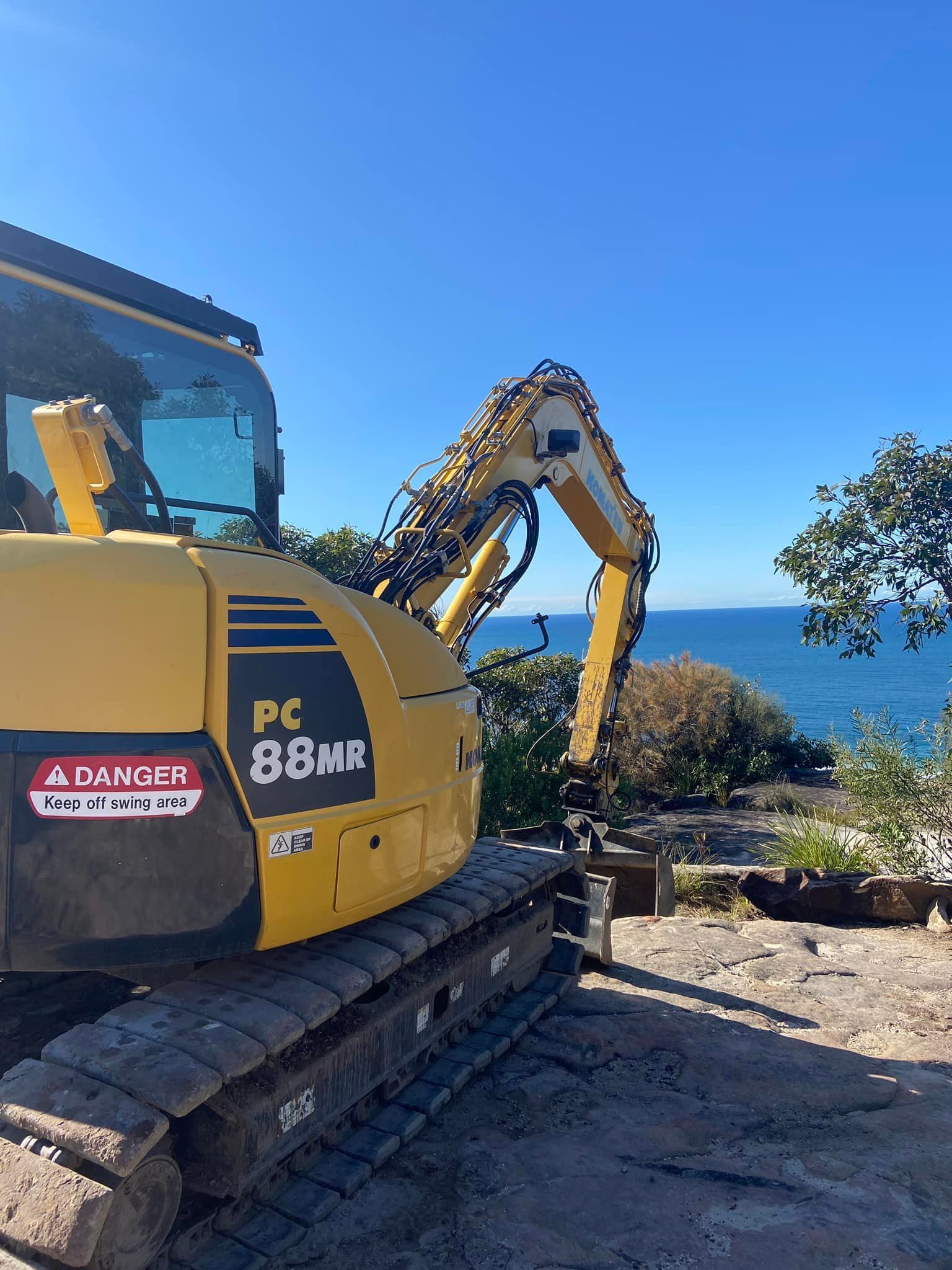 A yellow excavator is parked on a dirt road next to the ocean. — Adam Lowe Earthmoving Pty Ltd in Somersby, NSW