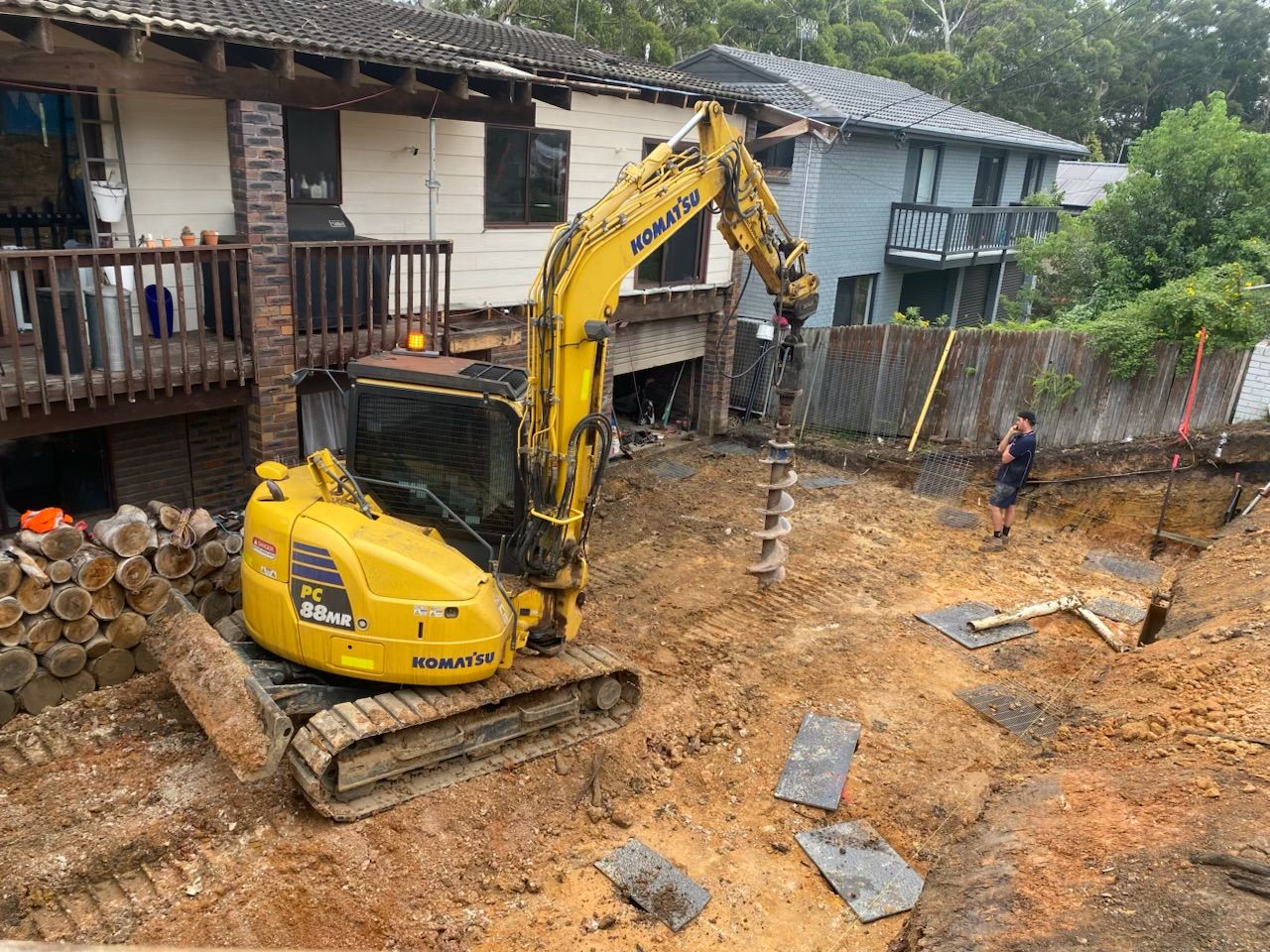 A yellow excavator is digging a hole in the dirt in front of a house. — Adam Lowe Earthmoving Pty Ltd in Somersby, NSW