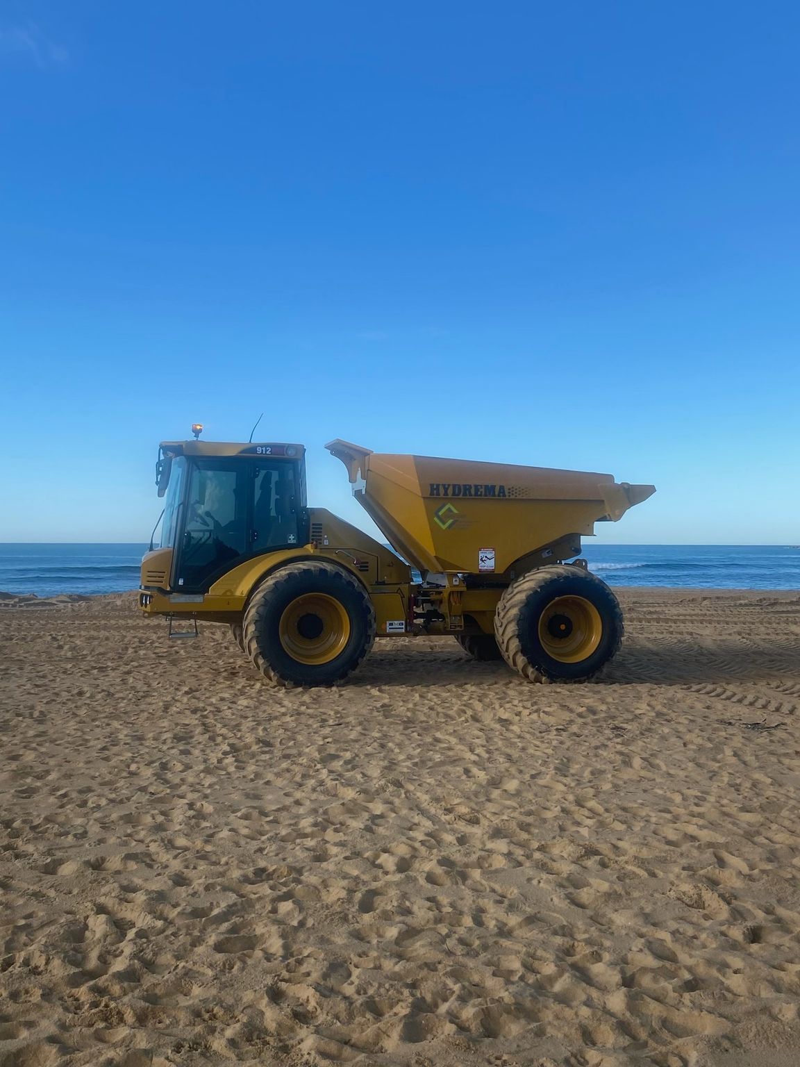 A yellow dump truck is parked on a sandy beach near the ocean. — Adam Lowe Earthmoving Pty Ltd in Somersby, NSW
