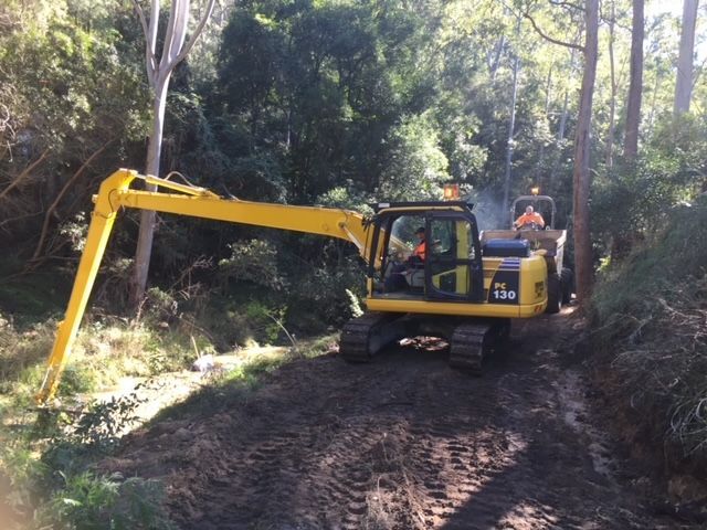 A yellow excavator is driving down a dirt road in the woods — Adam Lowe Earthmoving Pty Ltd in Somersby, NSW