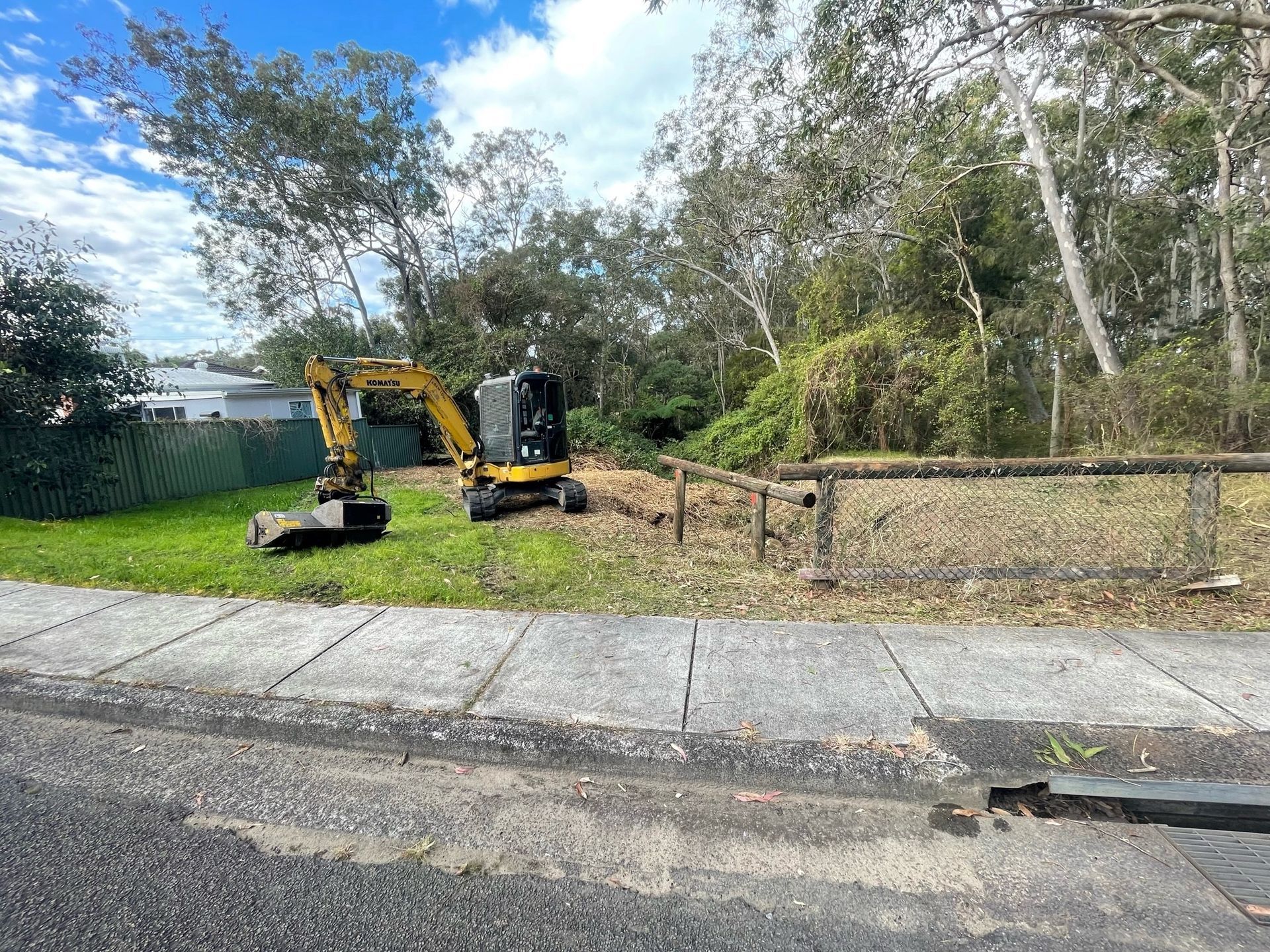 A yellow excavator is sitting in a grassy field next to a sidewalk. — Adam Lowe Earthmoving Pty Ltd in Somersby, NSW