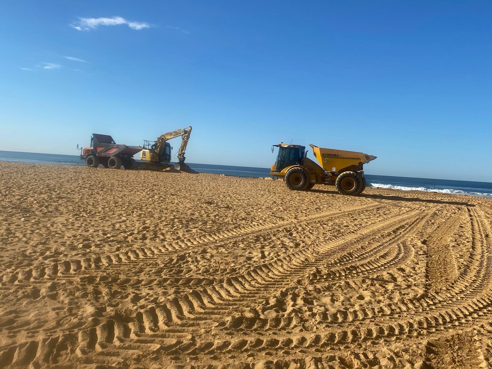 A dump truck is driving on a sandy beach next to a tractor. — Adam Lowe Earthmoving Pty Ltd in Somersby, NSW