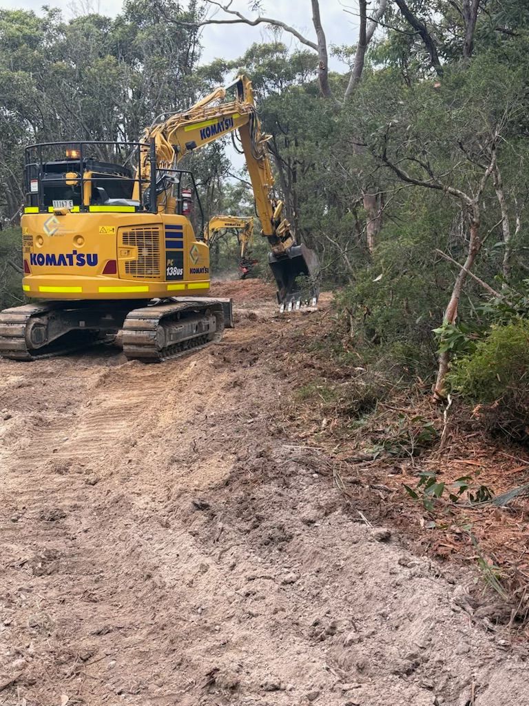 A yellow excavator is driving down a dirt road in the woods. — Adam Lowe Earthmoving Pty Ltd in Somersby, NSW