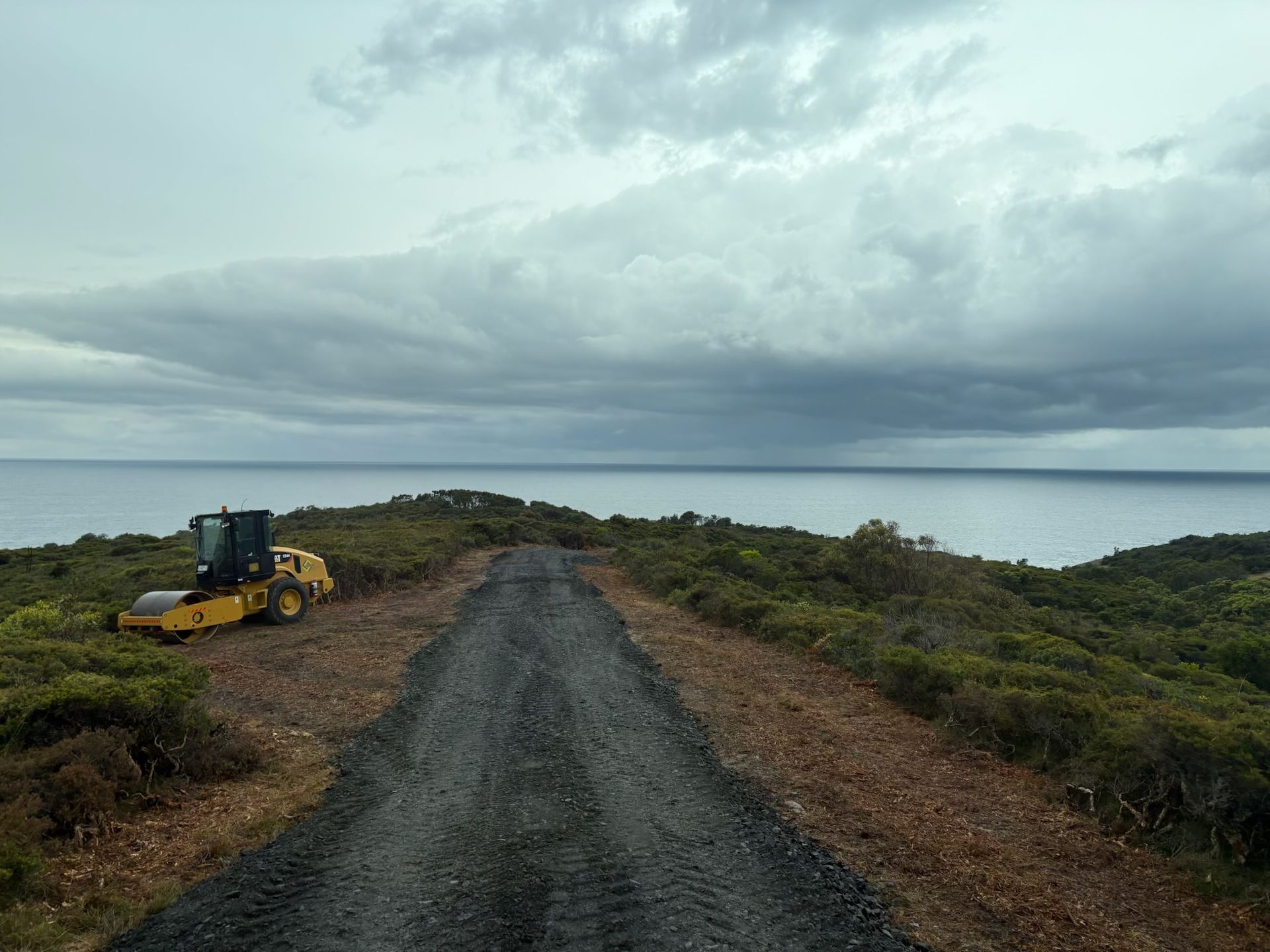 A yellow tractor is driving down a dirt road next to the ocean. — Adam Lowe Earthmoving Pty Ltd in Somersby, NSW